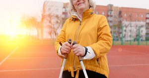 Vanhempi nainen seisoo parkkipaikalla keltainen tuulipuvun takki päällään sekä kävelysauvat käsissään.